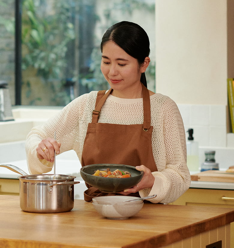 キッチンでエプロン姿の女性が鍋をかき混ぜ、シチューの入った器を持っている写真